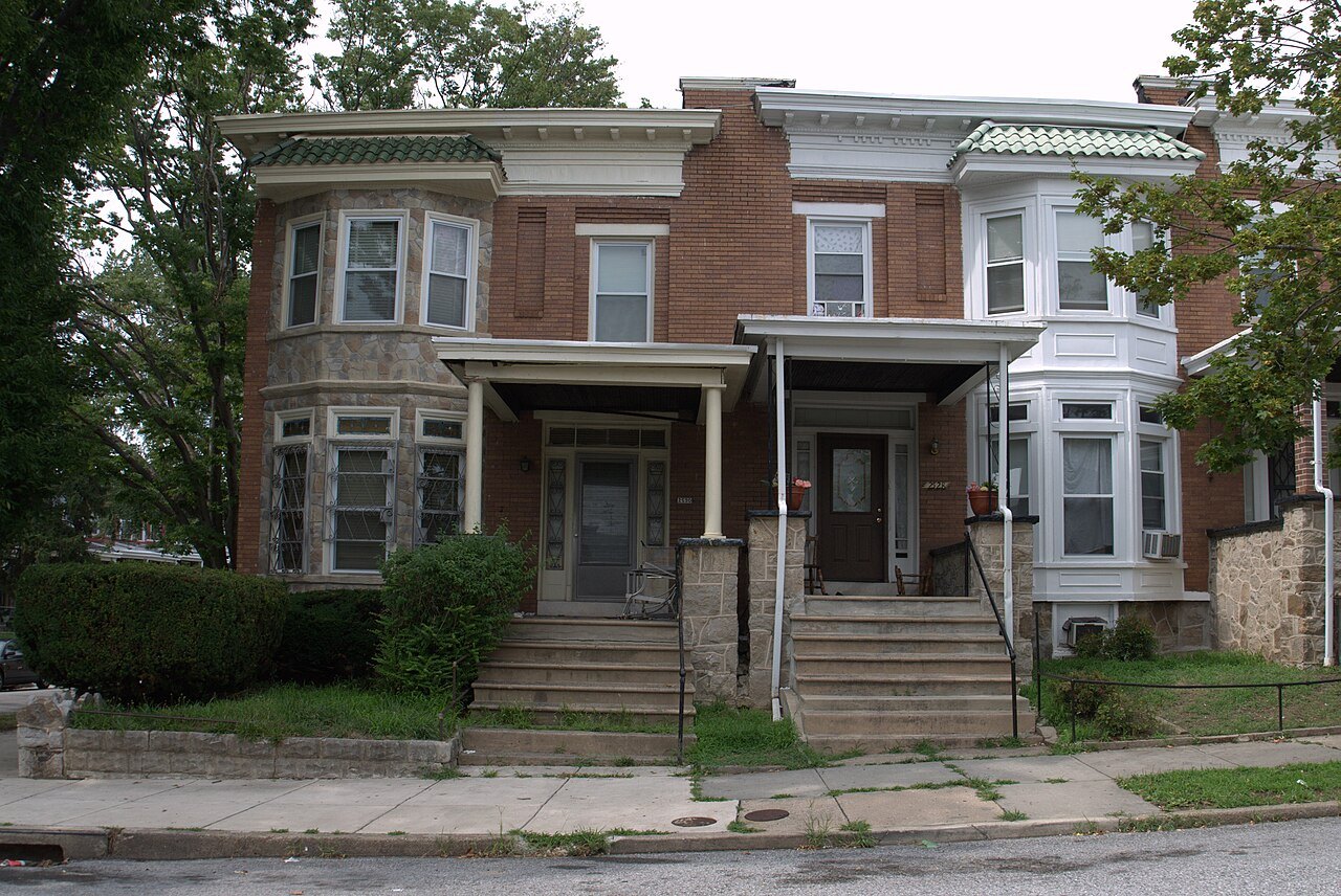 A row of brick and stone rowhouses with front porches and steps along a street in Baltimore