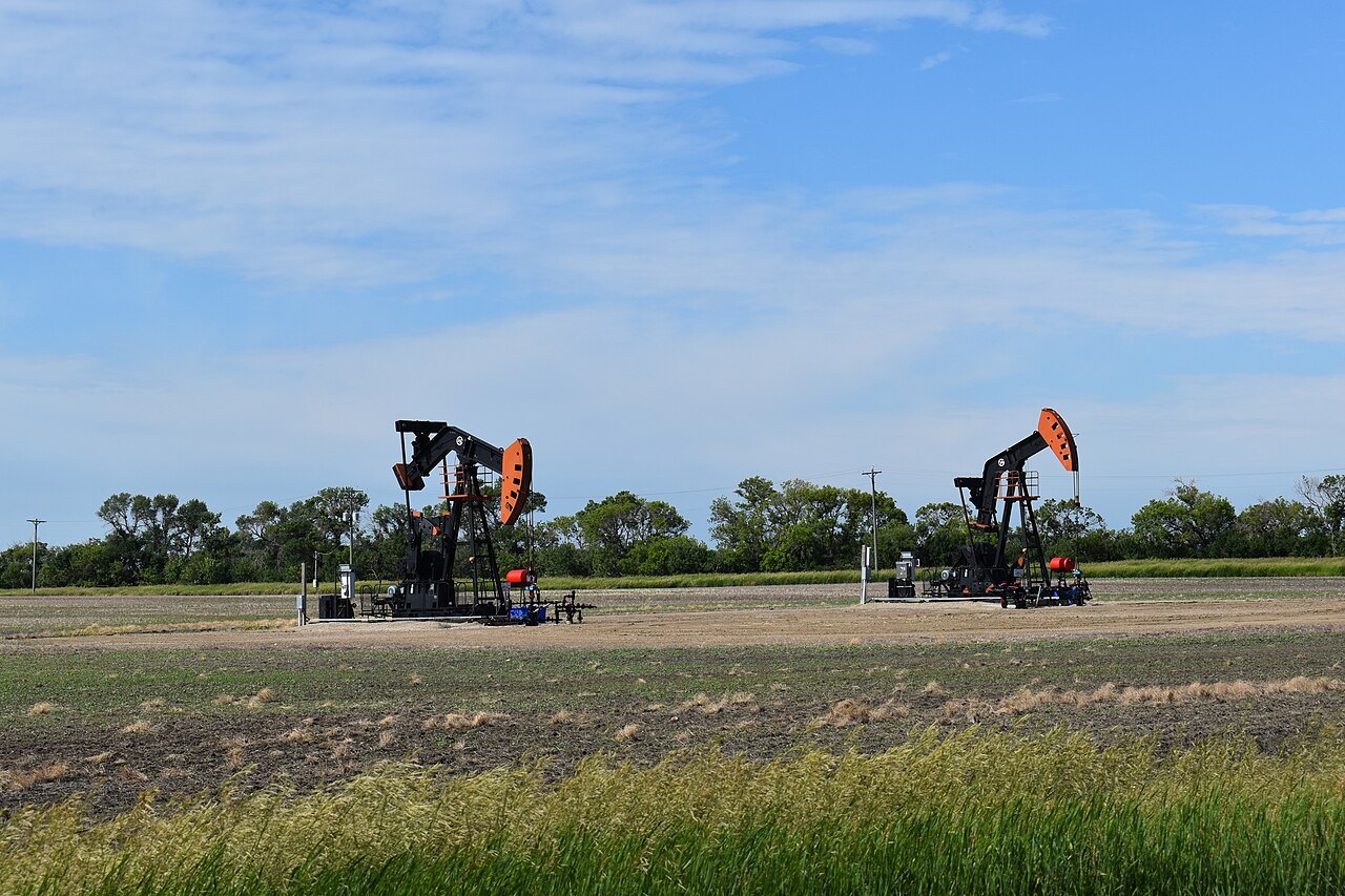 Two black and orange oil pumpjacks stand in a flat field with trees behind them under a blue sky