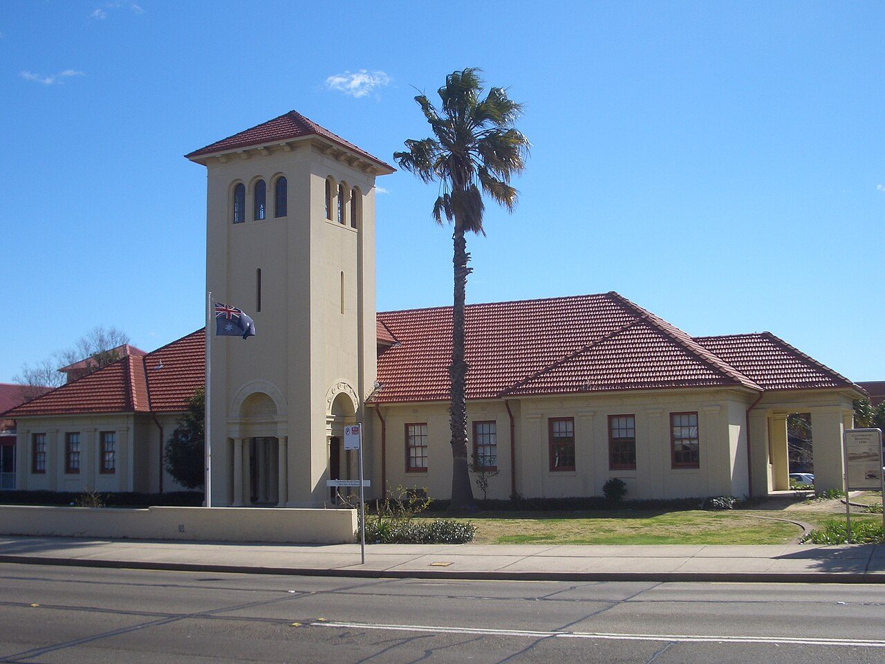 A cream hospital building with a red tiled roof and a square clock tower, next to a palm tree under a blue sky