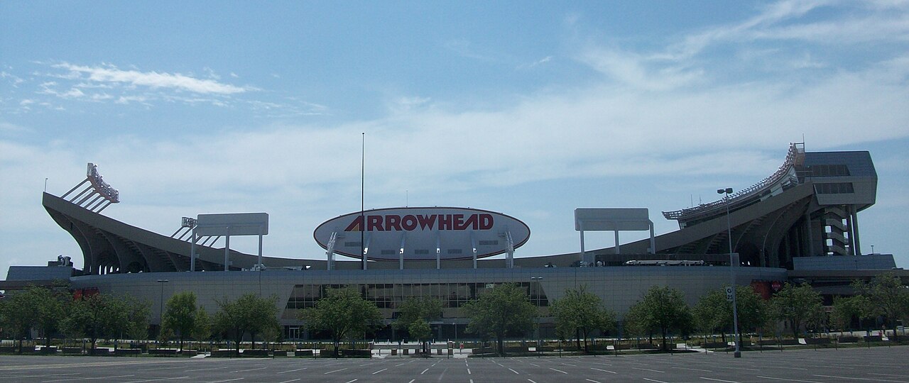 The curved red-trimmed roof and outer wall of a large football stadium under a blue sky with light clouds