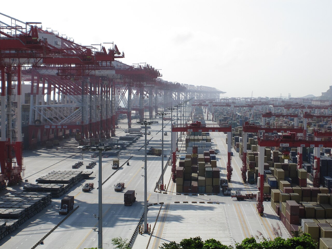 Rows of stacked shipping containers and tall red cranes at a large container port under a pale sky