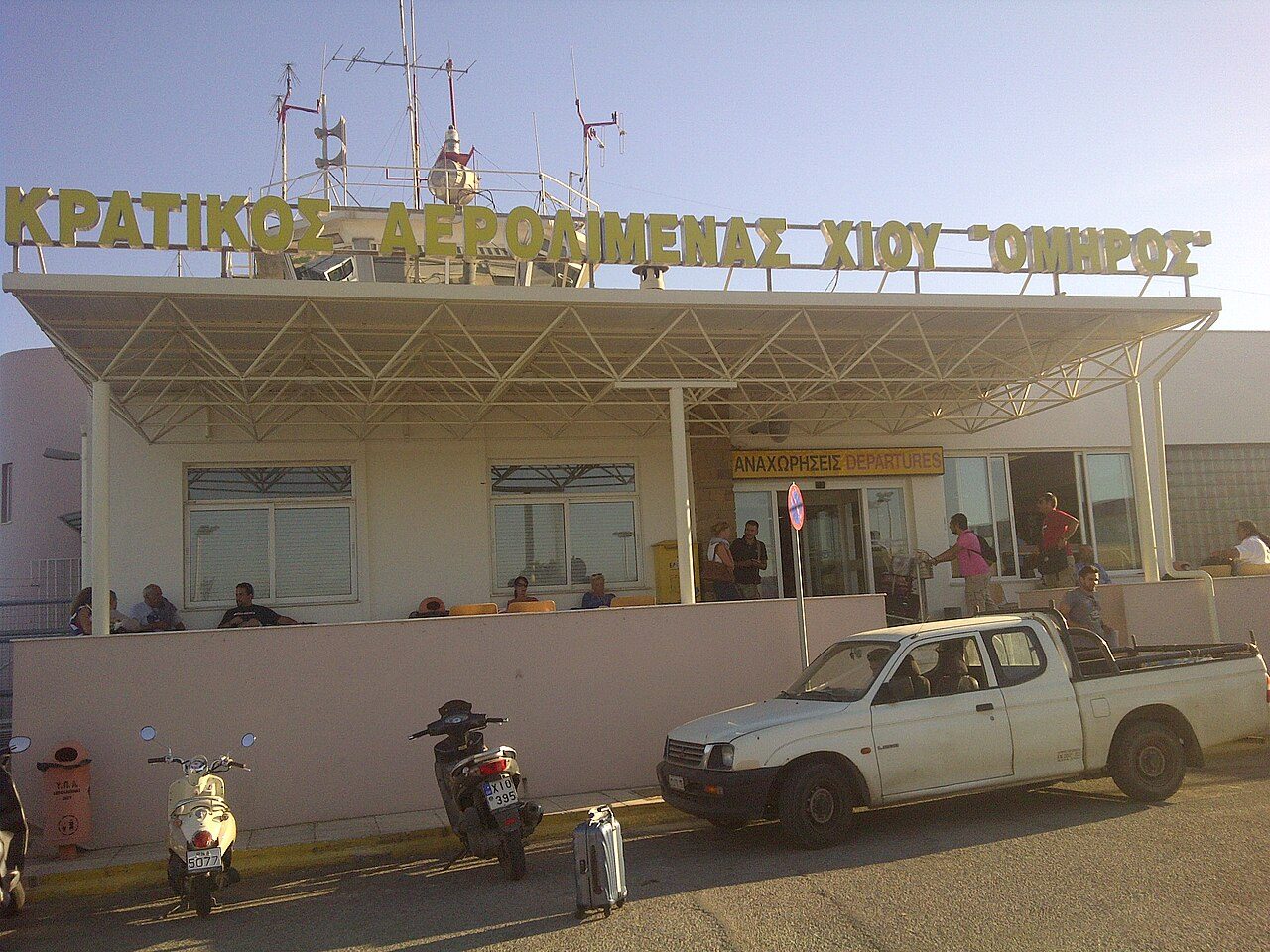 The front of a small airport terminal with a flat canopy and a sign in Greek letters, with a white pickup truck and parked motorbikes outside under a blue sky