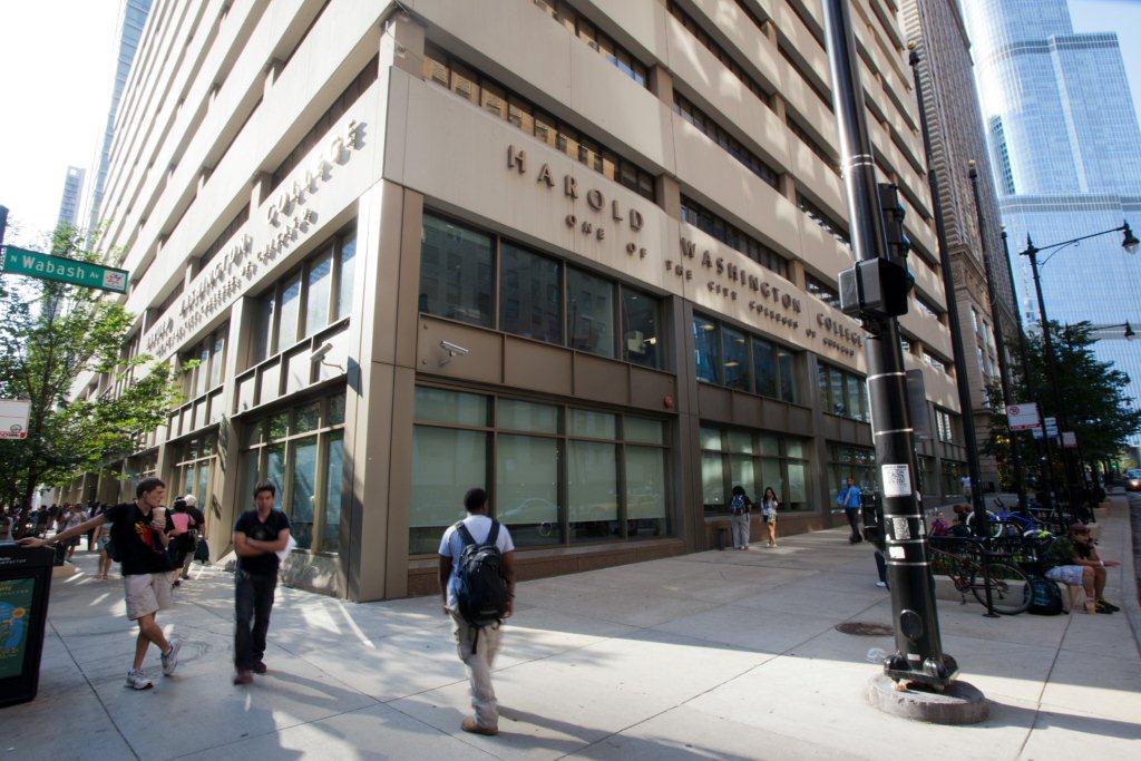 A tall stone-and-glass college building on a city street corner with people walking on the sidewalk in front of it