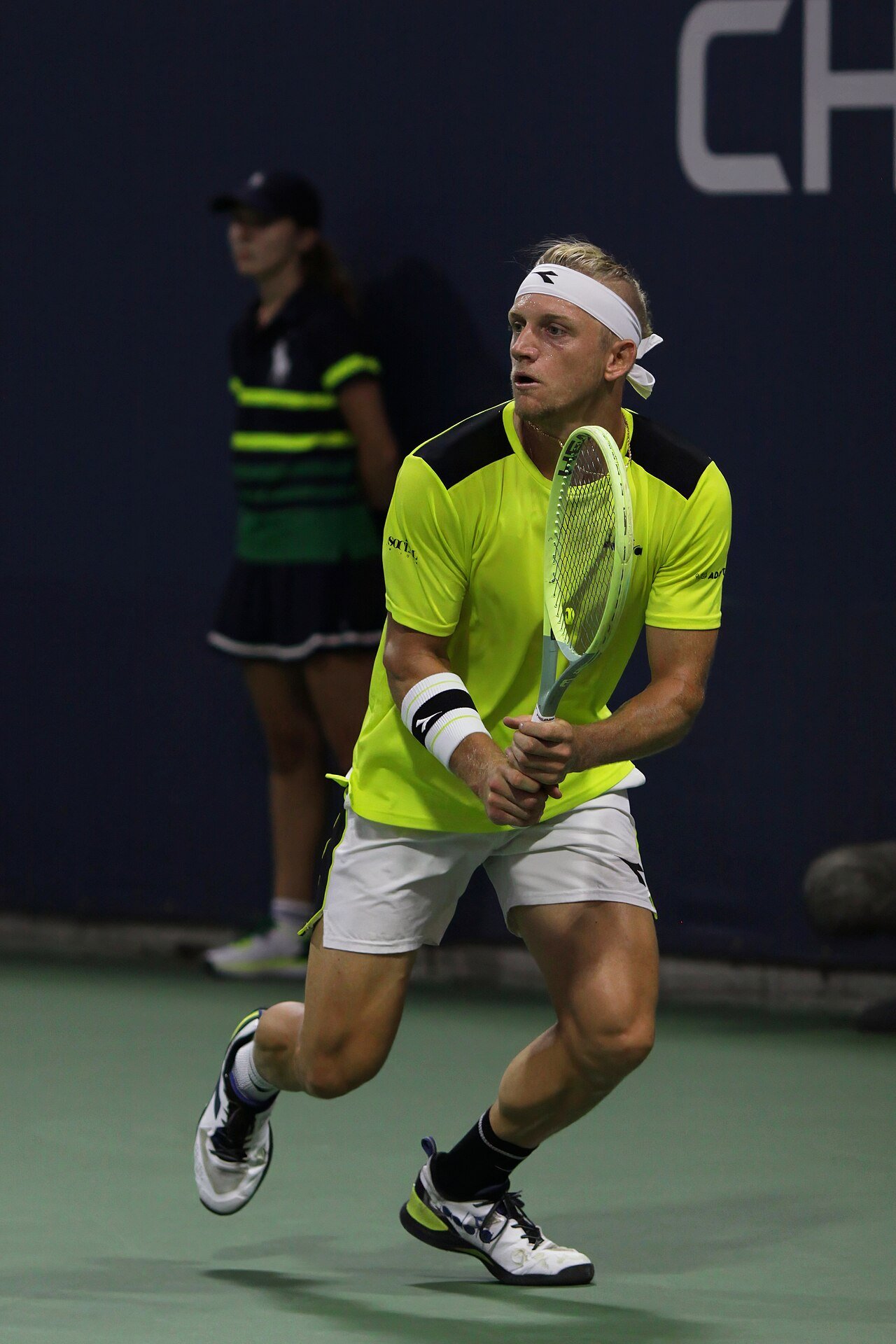 A tennis player in a bright yellow shirt and white headband runs toward the ball on a green hard court as a line judge stands behind him