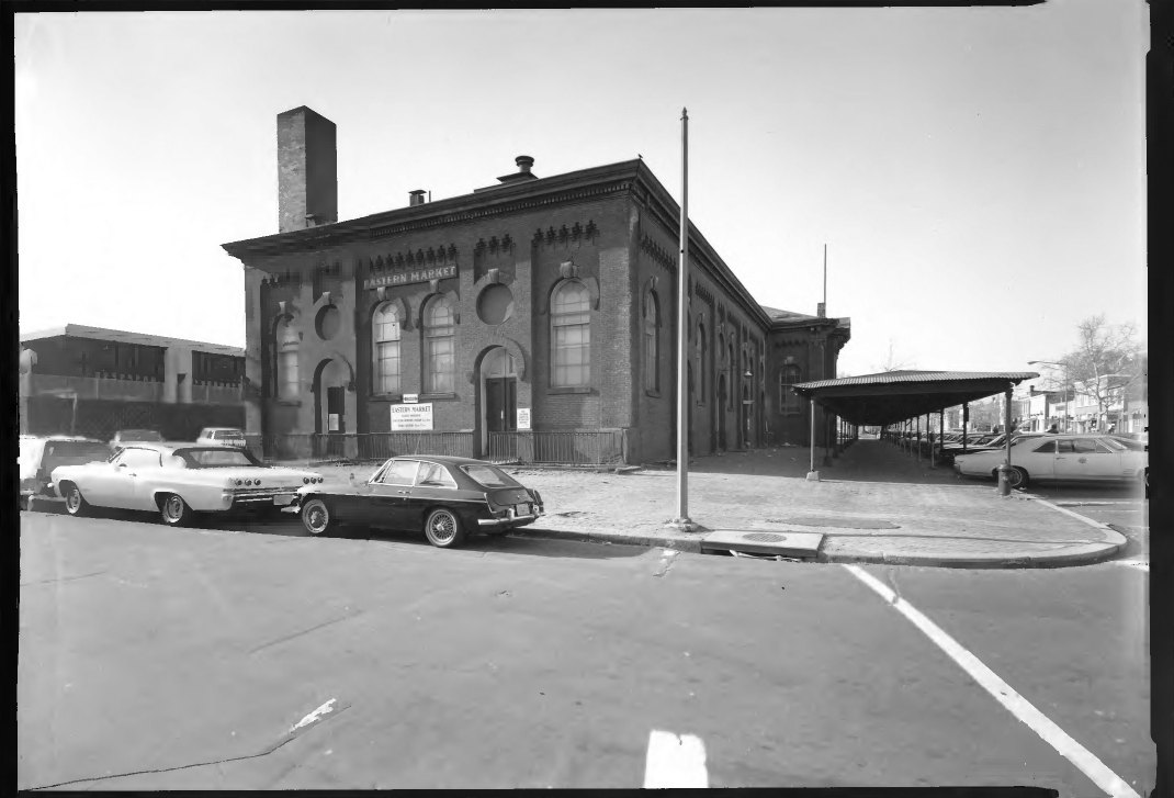 A black-and-white photo of the brick Eastern Market building in Washington, with cars parked out front