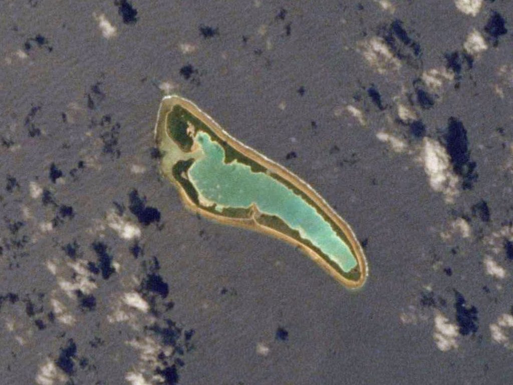 An aerial view of a narrow, ring-shaped island with a green lagoon in the middle, surrounded by dark blue ocean and scattered clouds