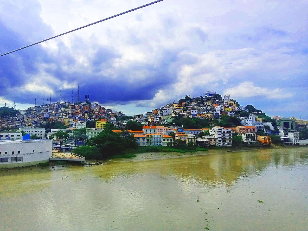 Colorful houses climb two green hills beside a wide brown river under a cloudy sky