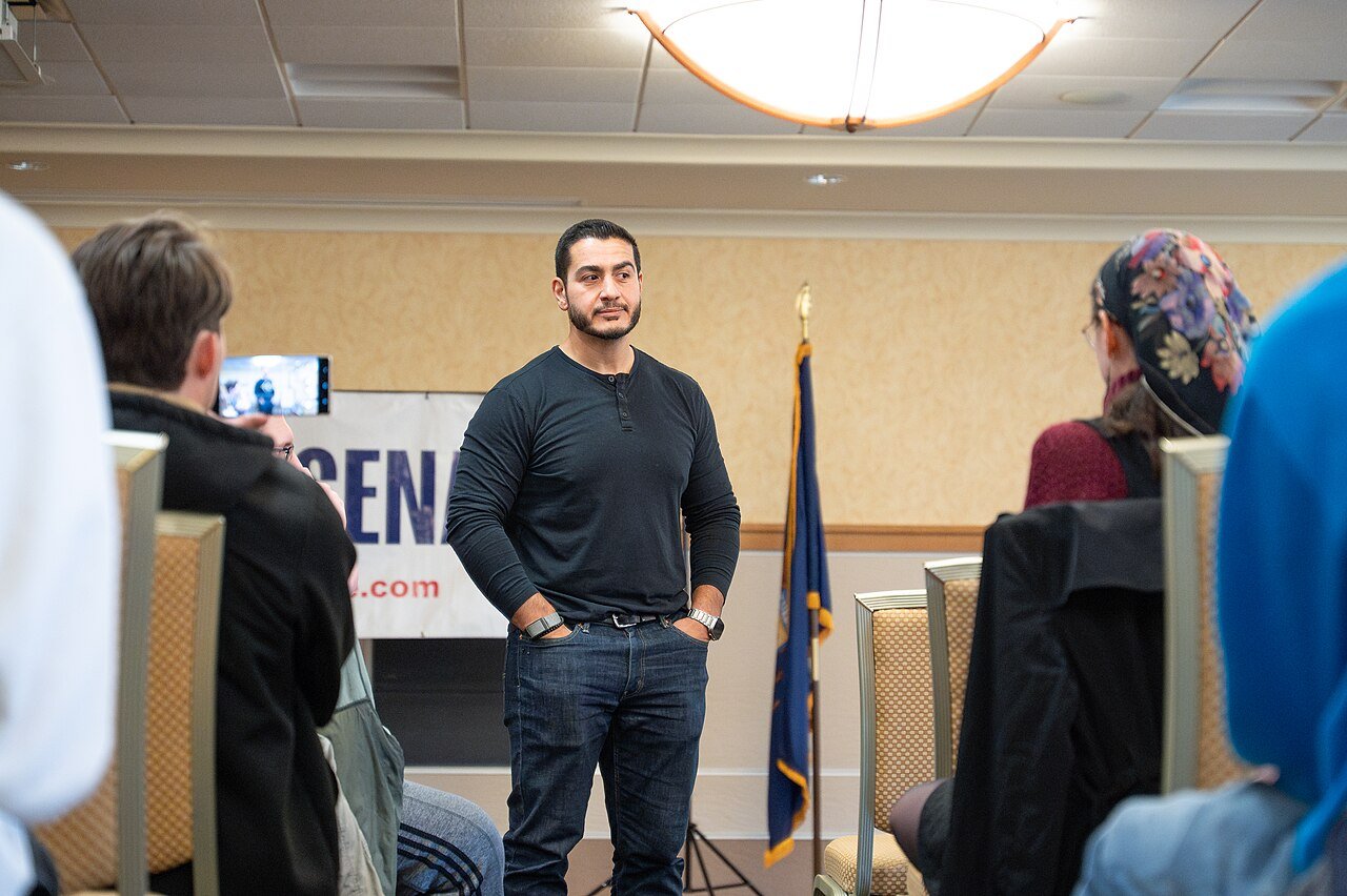 A man in a dark sweater stands speaking to a seated audience in a meeting room, with a campaign sign behind him