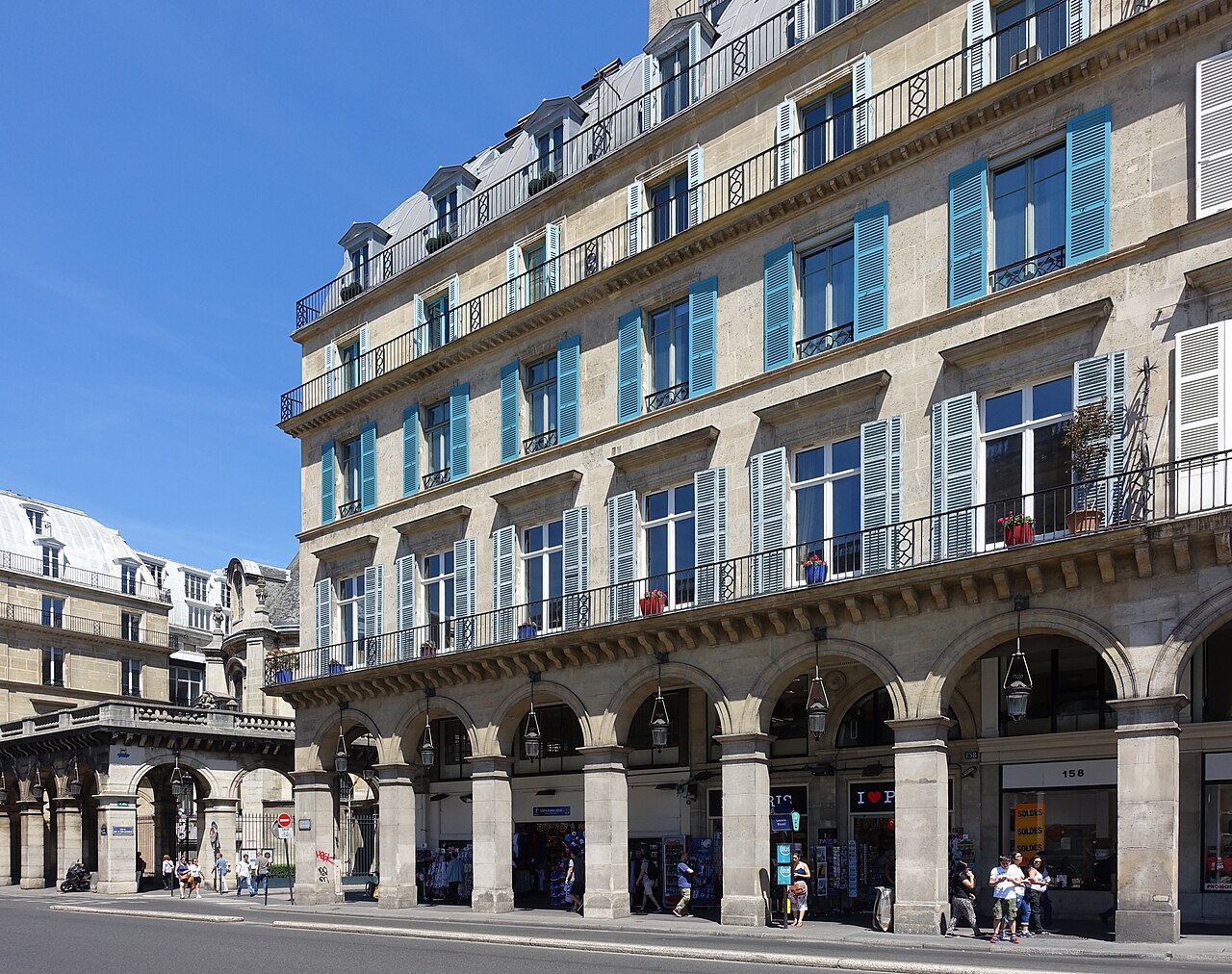 A row of Paris shopfronts under stone arcades on a sunny day, with people walking past