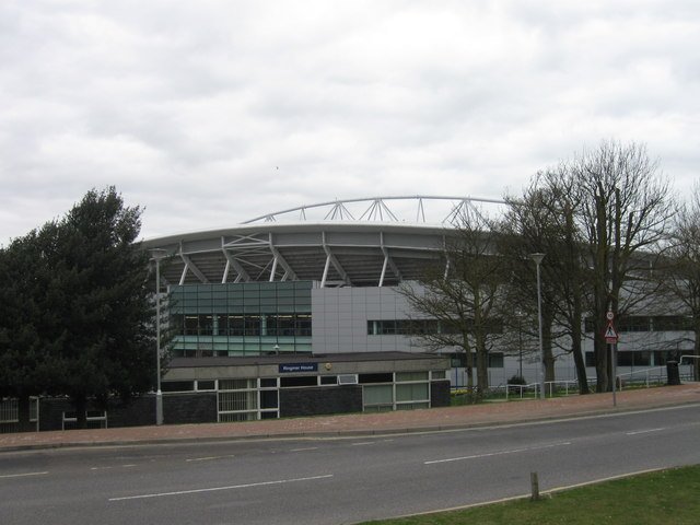 The curved white roof of the Amex Stadium in Brighton seen from a road lined with bare trees under a gray sky