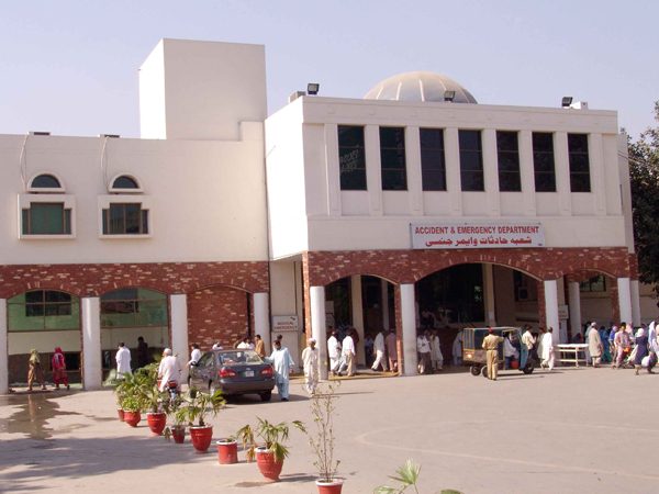 A white hospital building with an accident and emergency department sign over brick arches, with people and a car outside