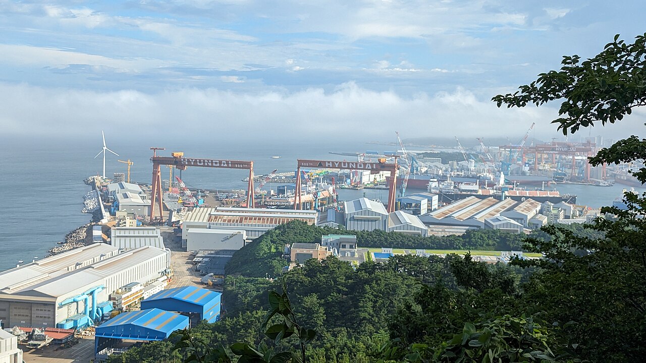 A coastal shipyard with red gantry cranes and large buildings beside the sea, seen from a wooded hill