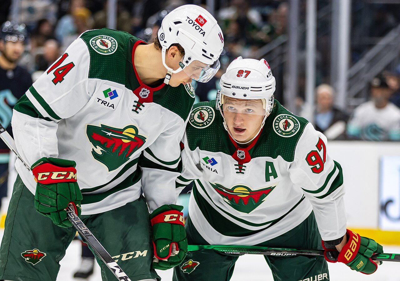Two Minnesota Wild players in white and green jerseys, numbers 14 and 97, lean in close on the ice during a game