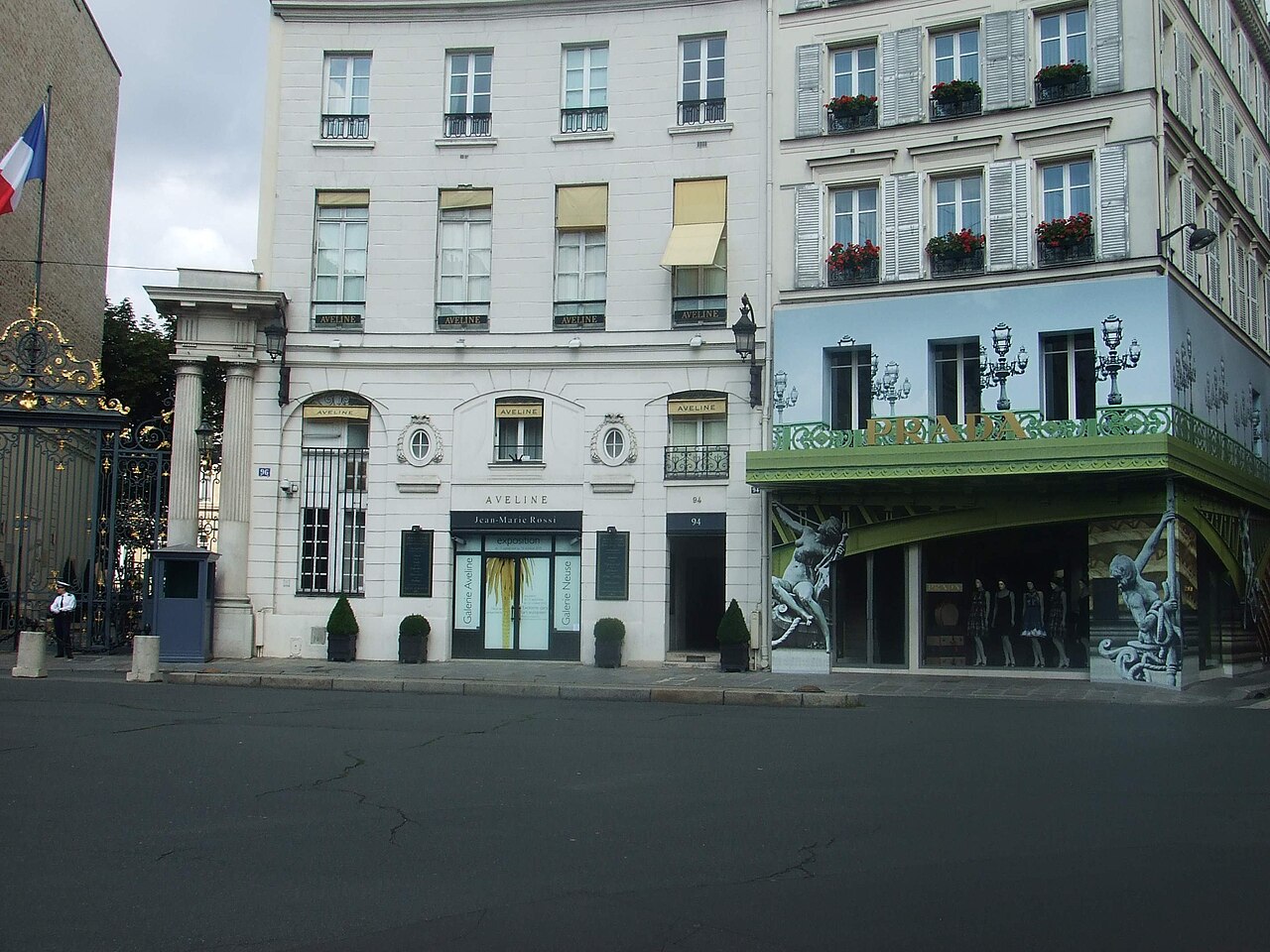 Stone apartment buildings with shops at street level and a French flag at the left edge, seen across an empty square