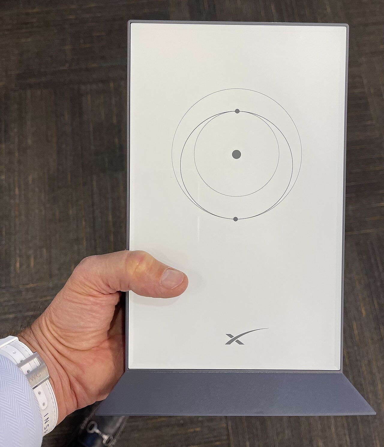 A hand holds a flat white rectangular satellite internet terminal on a grey stand, with an orbit diagram and the SpaceX logo on its face
