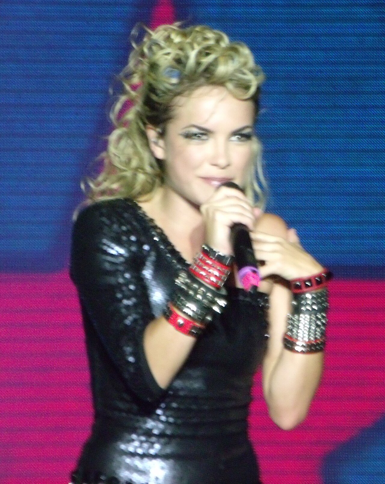 A woman with curly blond hair, wearing a black sequined top and studded wristbands, sings into a microphone on a stage