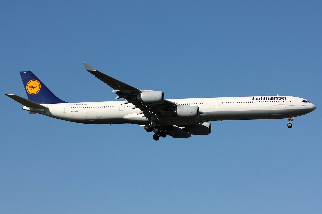 A white Lufthansa four-engine jet with a dark blue tail flies with its landing gear down against a clear blue sky