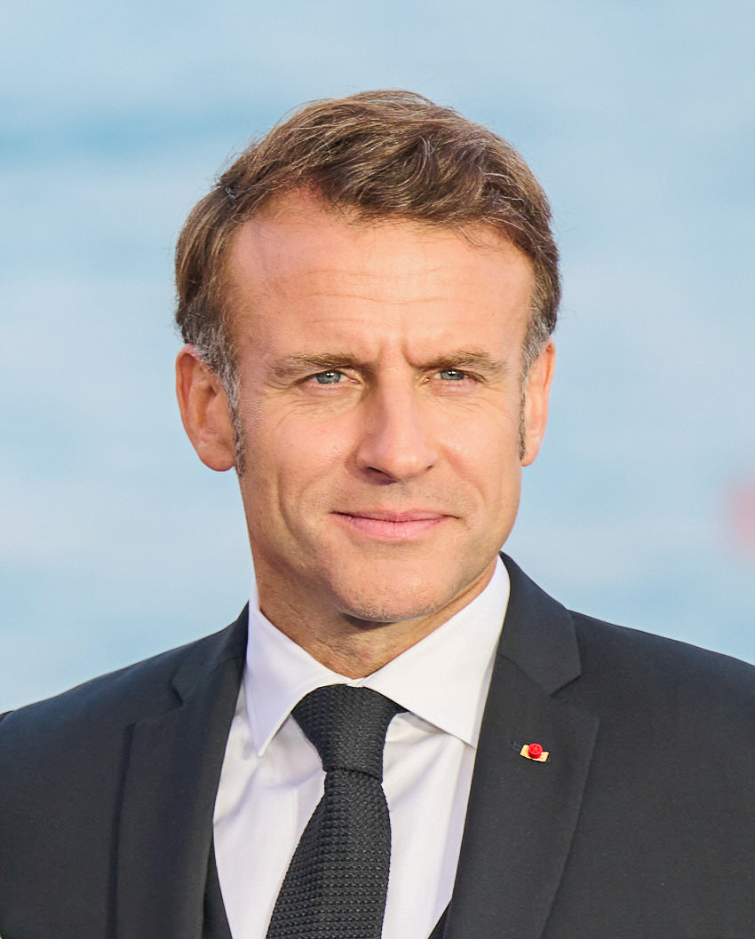 Emmanuel Macron in a dark suit, white shirt and dark tie, smiling slightly against a pale blue background