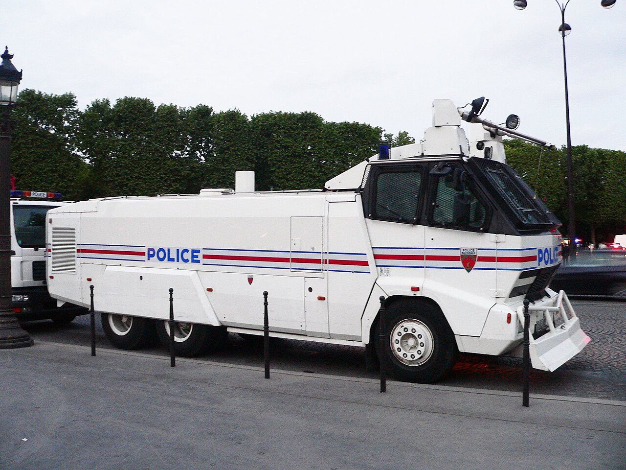 A white French police water cannon truck with a red and blue stripe and the word POLICE on its side is parked on a tree-lined street