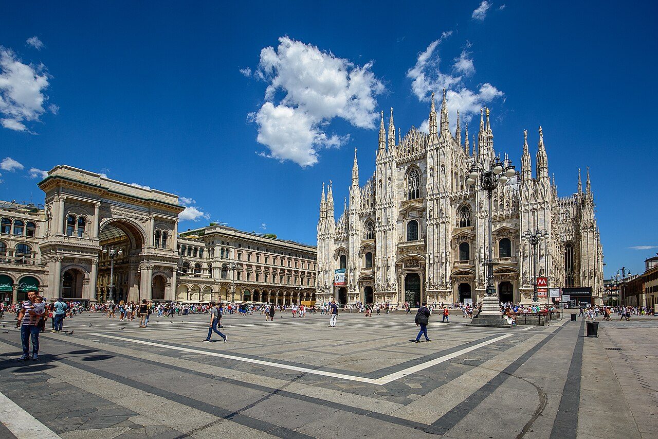 A wide, sunny city square with people walking across it, with the white Gothic facade of Milan Cathedral on the right and an arched gallery entrance on the left