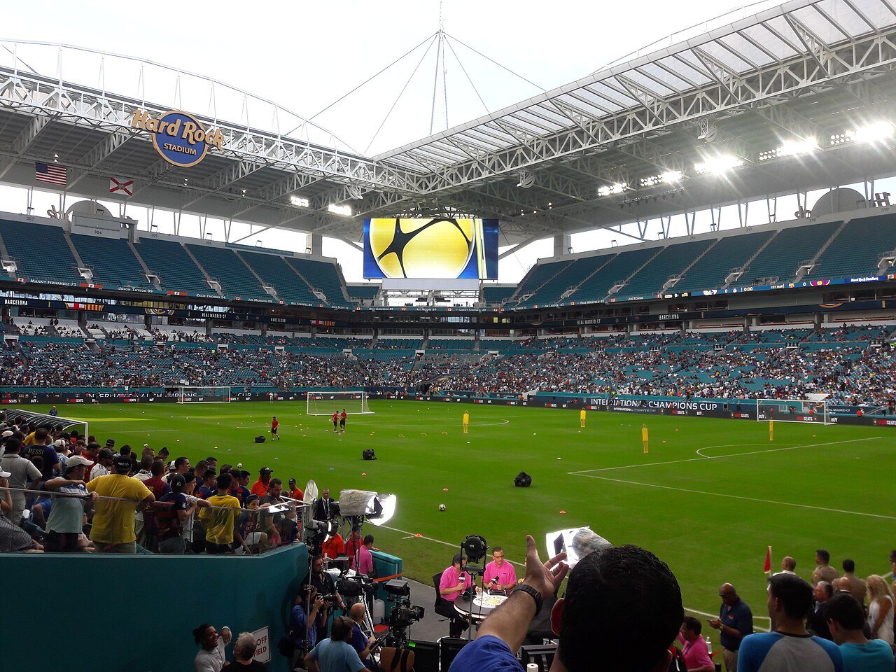 Inside a large stadium with a curved white canopy roof, crowds fill the stands around a green field during a match