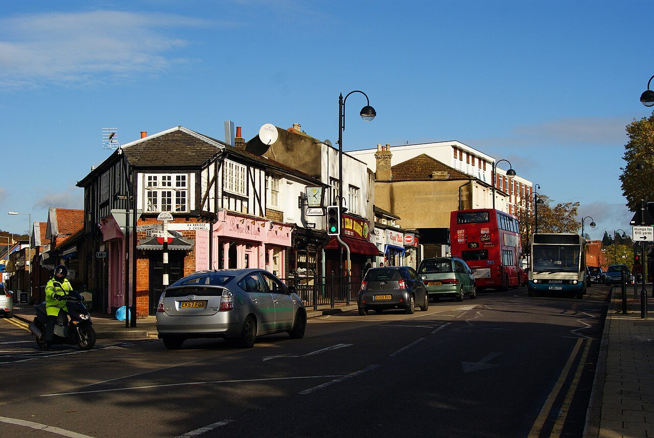 Cars and a red double-decker bus travel along a sunlit high street lined with shops and a black-and-white timber-framed building