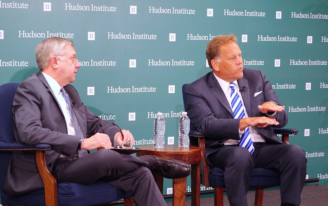 Two men in suits sit in armchairs on a stage in front of a teal backdrop, one speaking and gesturing with his hand