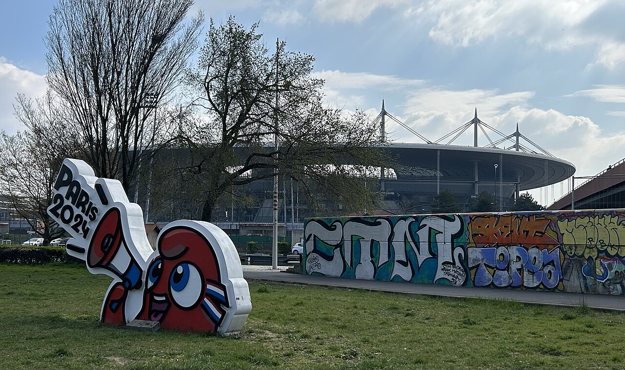 A large red, white and blue cartoon sculpture covered in graffiti stands on grass, with the roof of Stade de France behind it