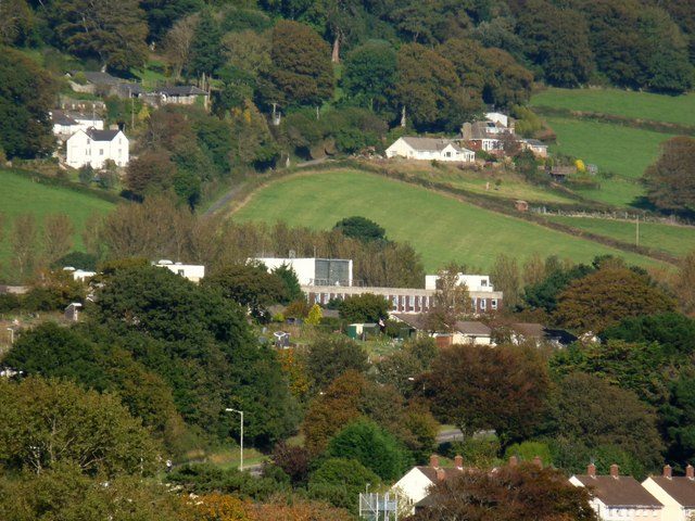 Low white hospital buildings among trees on a green hillside, with houses and fields above