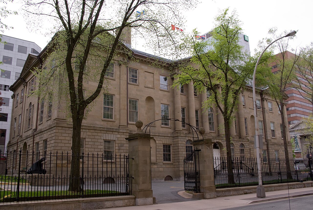 A three-story sandstone government building with tall windows stands behind an iron fence and leafy trees on a city street