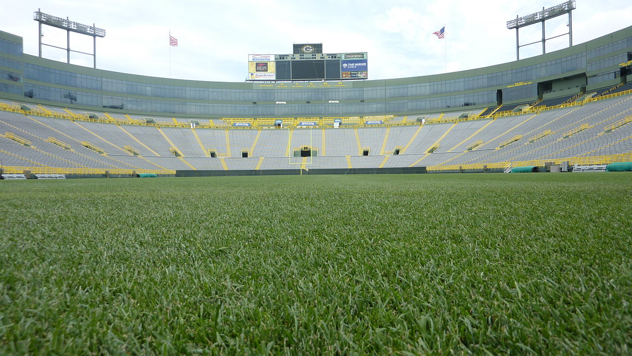 A green grass football field in a large stadium with yellow and green seats and a scoreboard at the far end