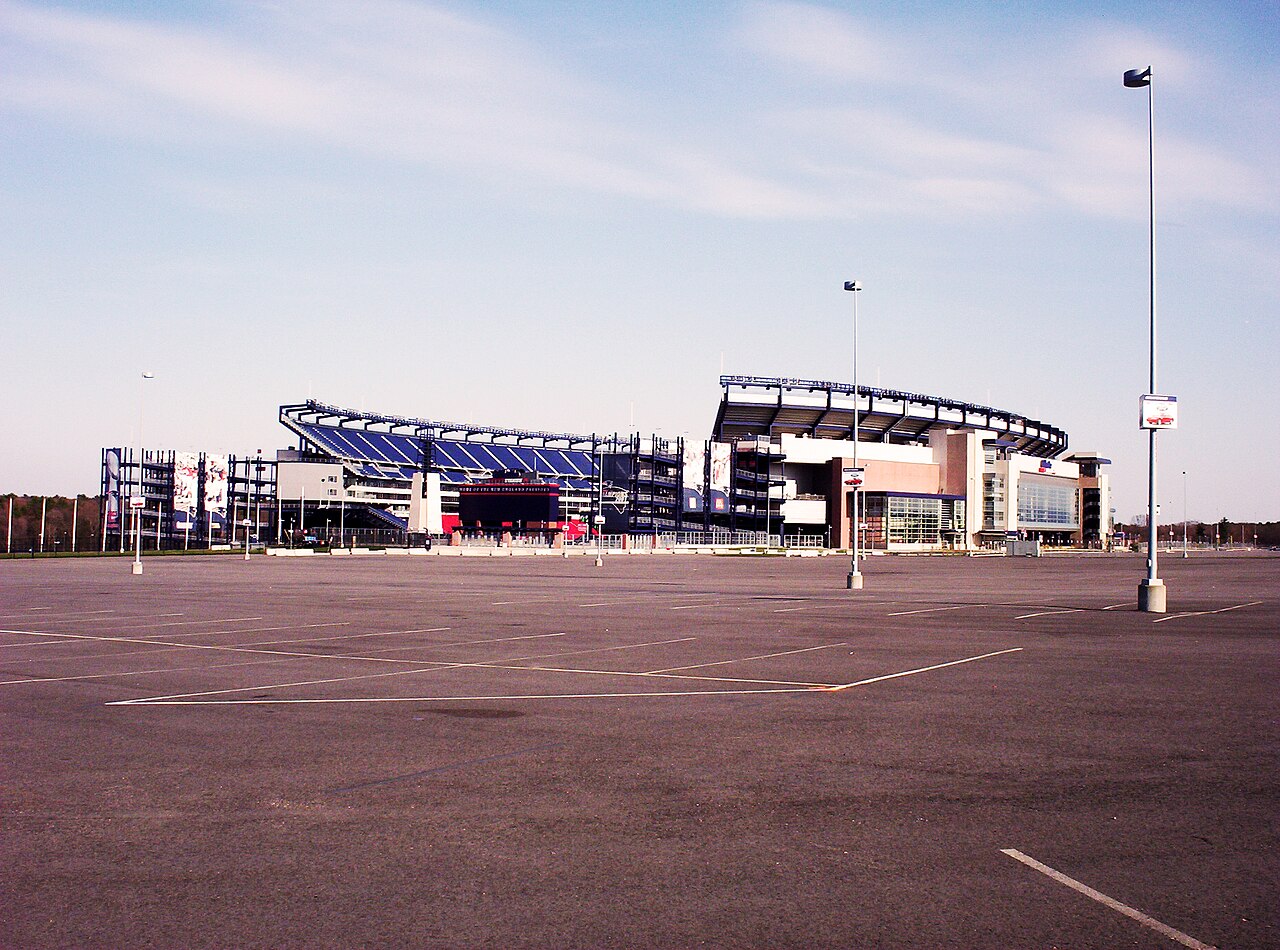 A large football stadium with blue seating stands behind an empty paved parking lot under a pale sky