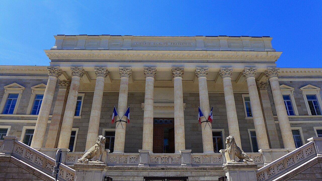 The columned stone front of a courthouse with flags above the entrance, under a blue sky