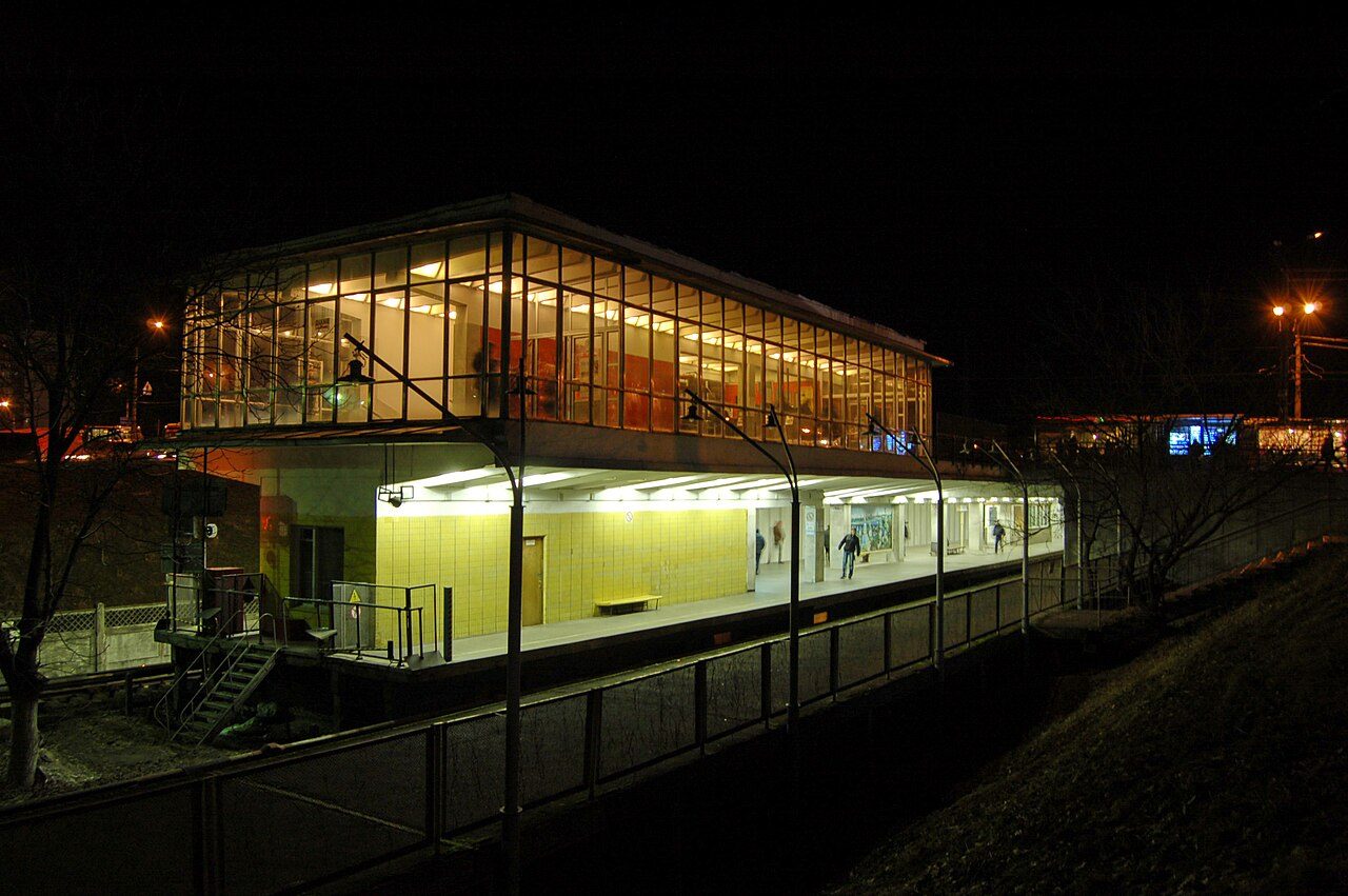 A lit, glass-walled metro station building stands on a raised platform at night, with a few people walking inside