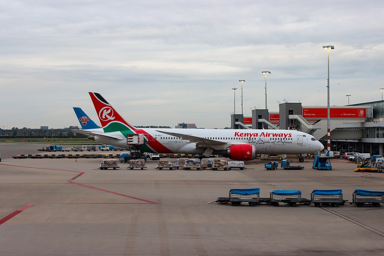 A red and white Kenya Airways jet parked at a gate at Amsterdam Airport Schiphol under a cloudy sky, with baggage carts in front