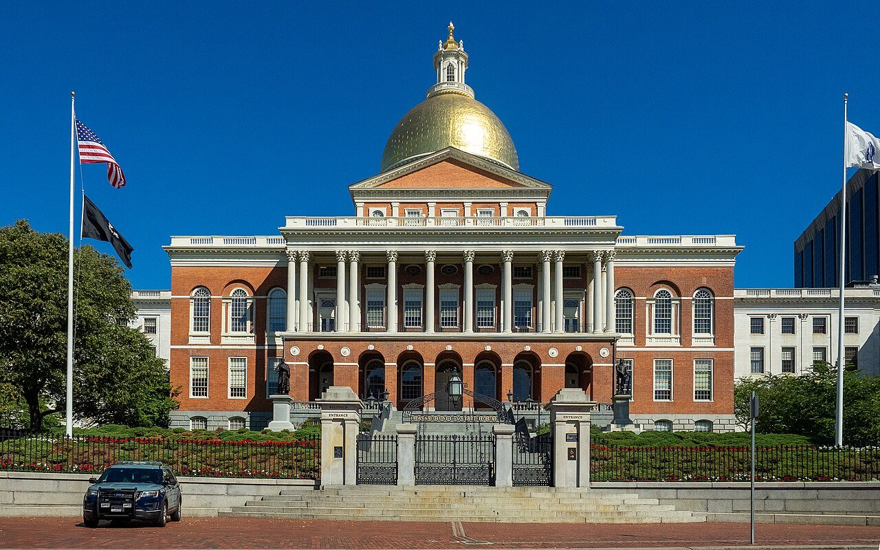 A red brick government building with white columns and a gold dome stands behind a gated lawn and flagpoles