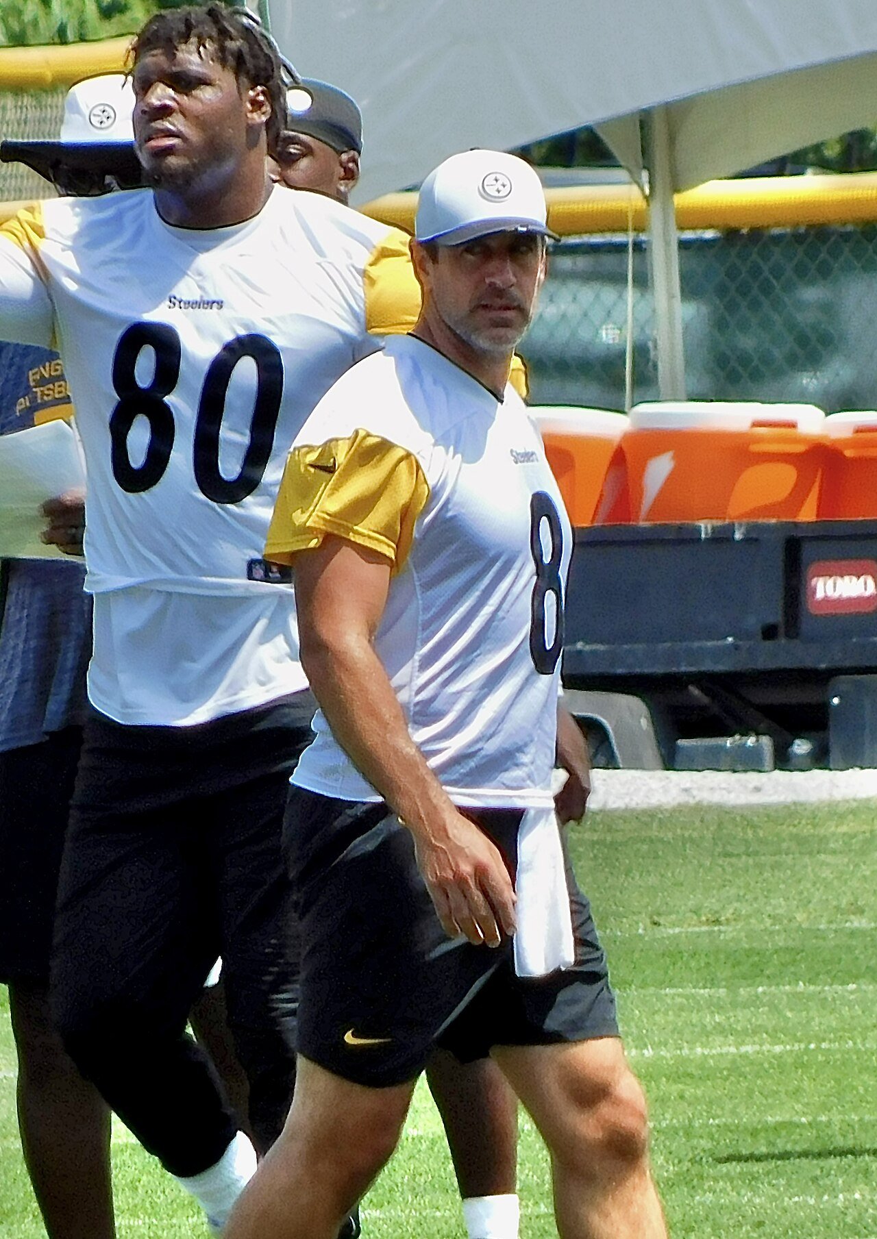 Aaron Rodgers in a white Steelers jersey and cap walks on a grass field next to a teammate wearing number 80