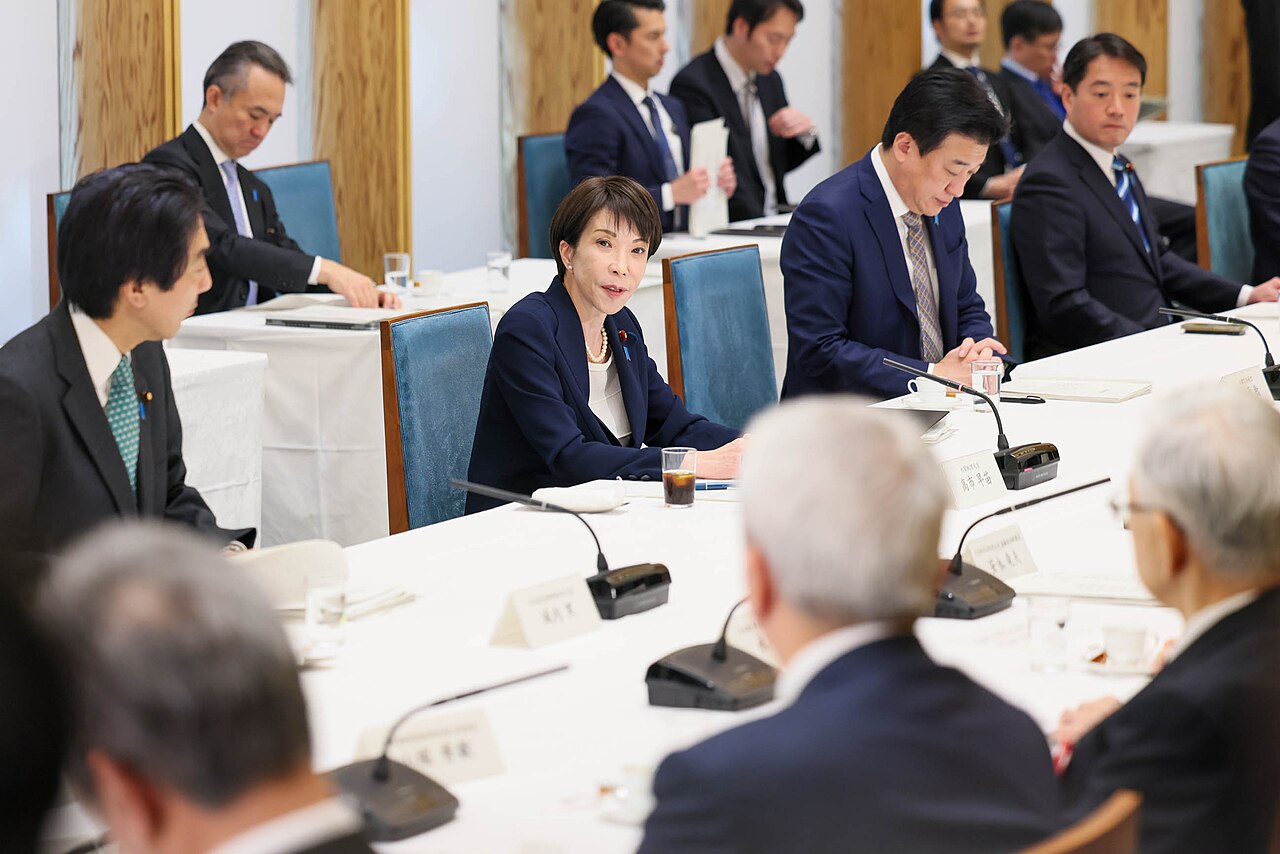 A woman in a dark jacket sits at a long white conference table with men in suits during a meeting in a wood-panelled room