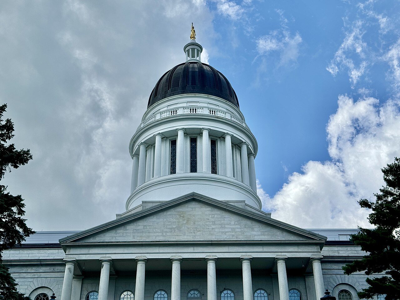The dome and columned front of the Maine State House stand under a blue sky with scattered clouds
