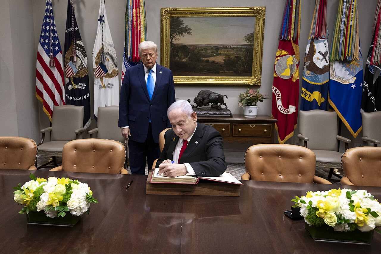 Donald Trump stands behind Benjamin Netanyahu, who sits at a wooden table signing a book in a White House room with flags behind them