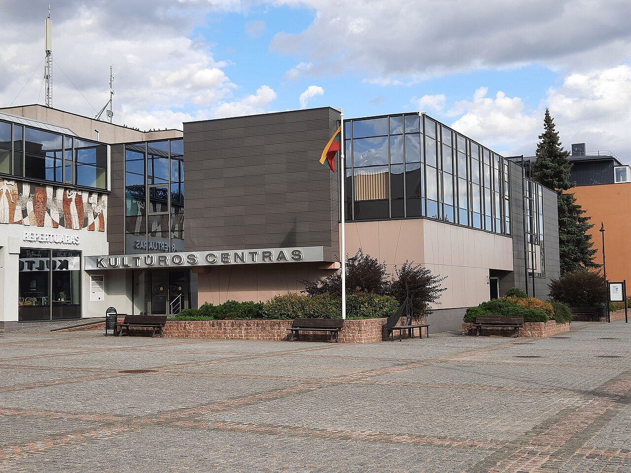 A modern two-story building with glass panels, a stone facade and flags out front stands beside a paved plaza with trees and flower beds under a blue sky