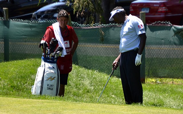A golfer in a white shirt and cap lines up a shot on grass beside a fence while a caddie in a red shirt stands by a golf bag
