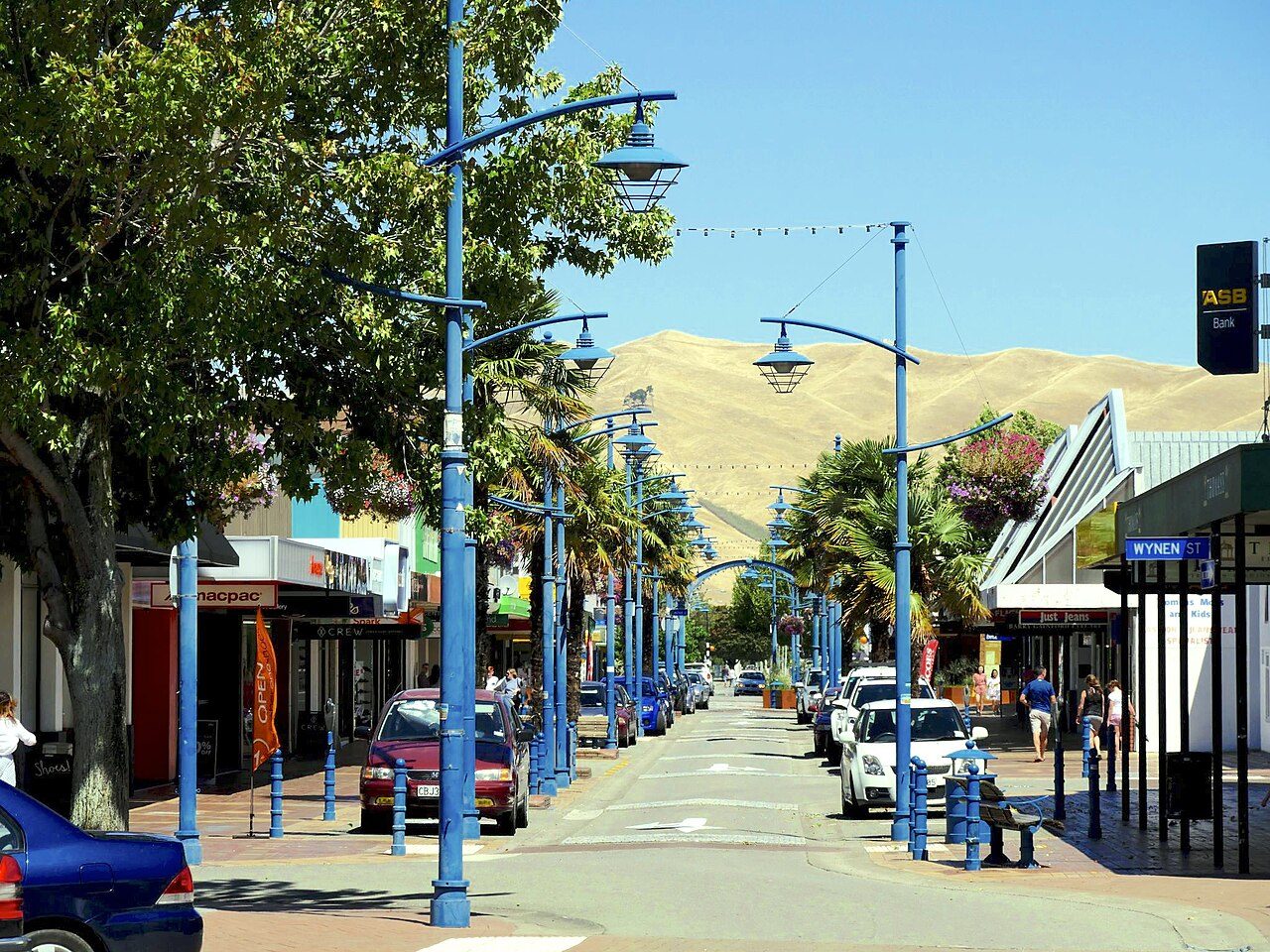 A sunny street lined with shops, blue lamp posts and parked cars, with hills in the background