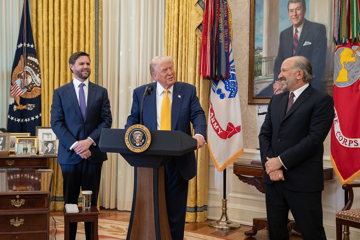 President Donald Trump speaks at a lectern in the Oval Office as Vice President JD Vance looks on