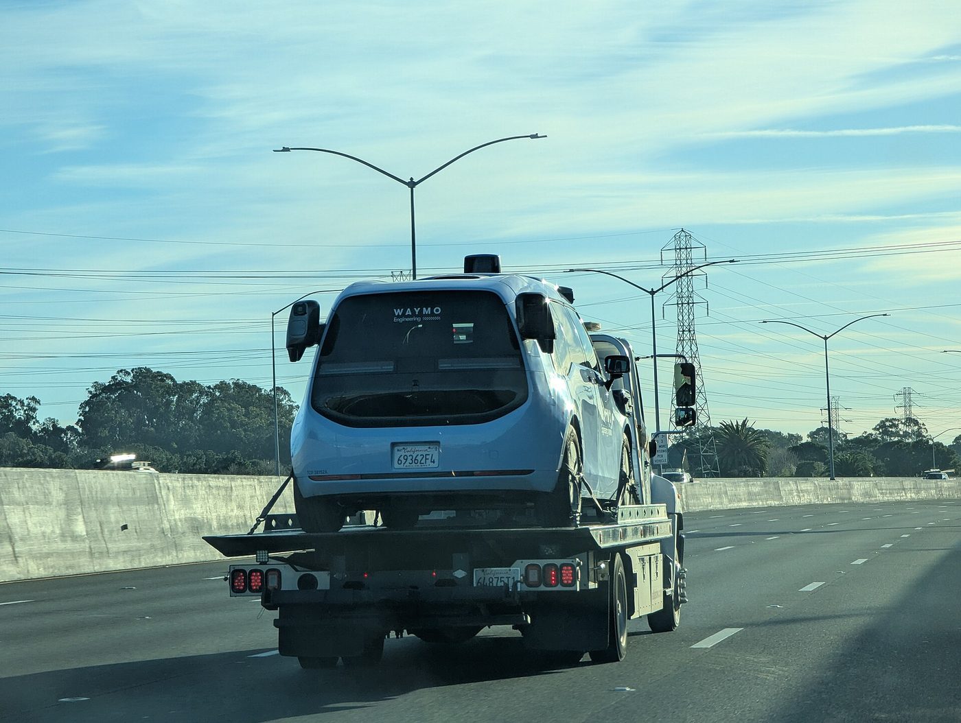 A white Waymo Ojai robotaxi van with roof sensors riding on a flatbed truck on a highway