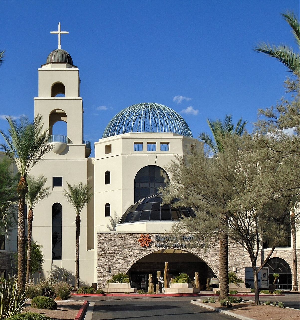 A hospital entrance with a bell tower topped by a cross and a blue dome, framed by palm trees