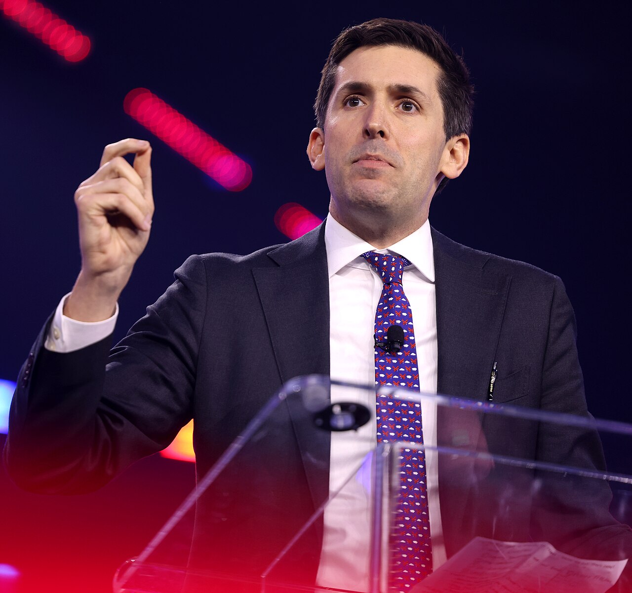 A man in a dark suit and purple tie speaks and gestures behind a clear lectern