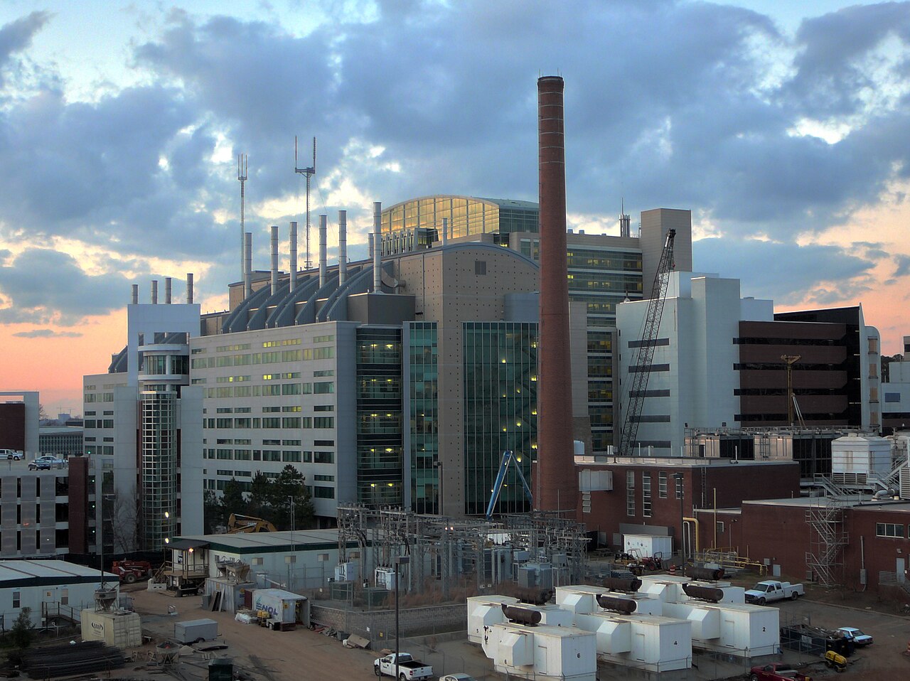 Modern office and lab buildings with a tall smokestack under a cloudy evening sky