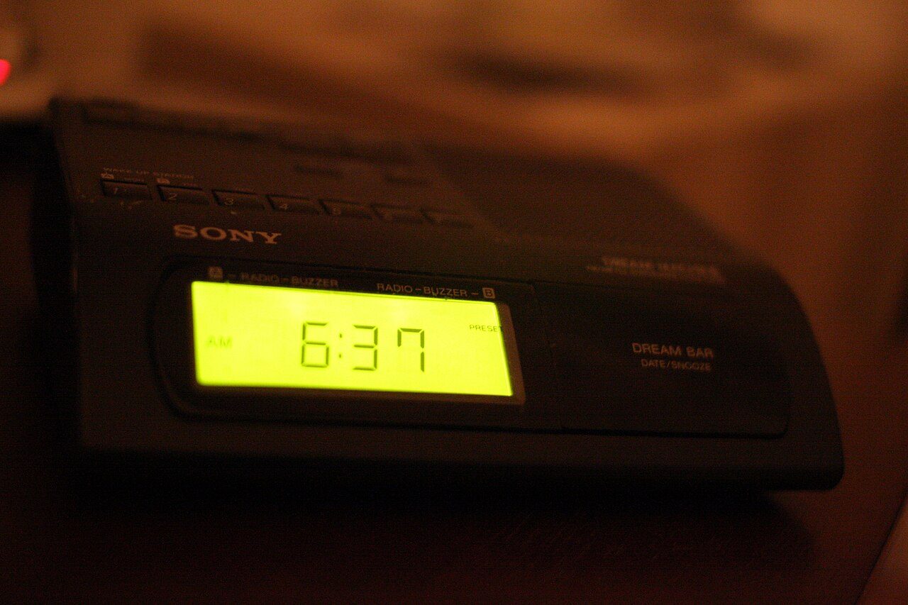 A black Sony clock radio on a bedside table glowing with the time 6:37 in a dark room