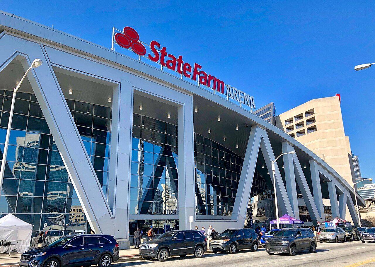 The glass and white steel front of State Farm Arena in Atlanta under a blue sky, with cars parked outside